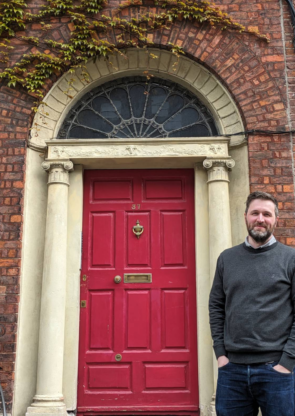 Ian wearing in a grey nit sweater over a button down shirt. Smiling in front of the red door of his office on North Great George Street